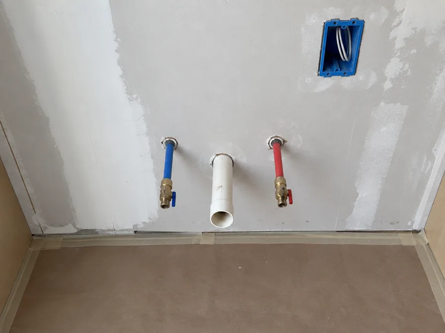 Bare drywall behind a removed base cabinet with new water supply stubs, a drain stub and a new outlet box