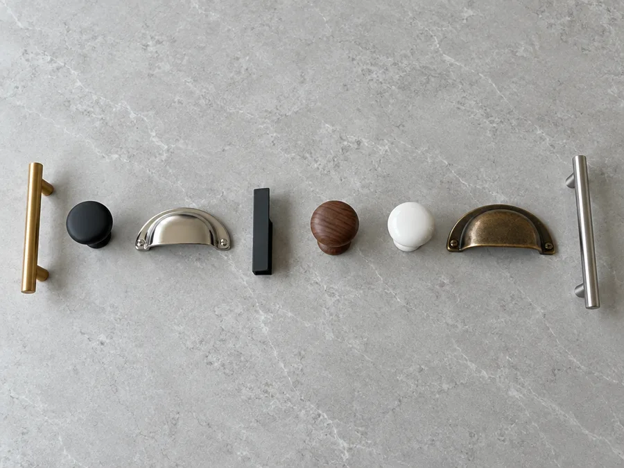 Eight different cabinet pulls and knobs in brass, black iron, nickel, wood and ceramic laid in a row on pale grey stone