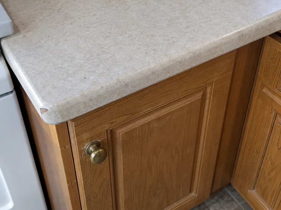 Close view of an older honey oak cabinet door below a worn laminate countertop edge with a chipped corner